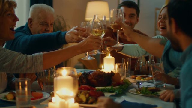 In the Evening: Family and Friends Gathered at the Dinner Table. Old and Young People Have Fun, Eating, Drinking. They Raise and Clink Glasses in a Toast. Cozy Living Room Atmosphere.
