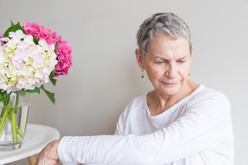 Portrait of beautiful older woman in white clothing looking down next to hydrangea flowers in vase (selective focus)