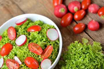 View from above of green salad Lollo Biondo with tomatoes and radishes in a white bowl on wooden table, a pitcher of oil in the background