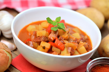 Potato and sausage goulash in bowl, pepper, onion and garlic on wooden table