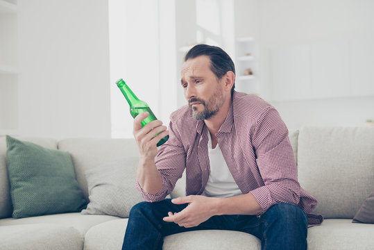 Man with sad face looking at the bottle sit on cosy couch in den