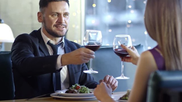 PAN of cheerful bearded man in suit and unrecognizable woman clinking glasses and drinking red wine on dinner date in restaurant