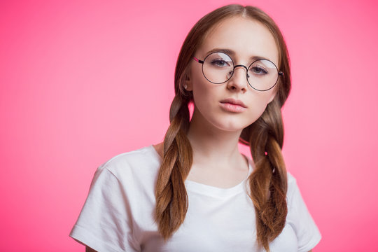 Close up portrait of young attractive girl with glasses and looking at camera on pink background. Redhead woman hands holding glasses