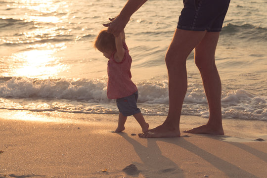 Father helping his baby with the first steps, teaching baby to walk concept, outdoor candid photo on the beach, spending a day at the beach, healthy family lifestyle.