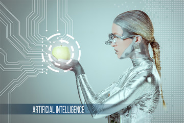 side view of cyborg holding and examining green apple with digital data isolated on grey with "artificial intelligence" lettering