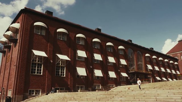 Woman in white jacket walking a dog down wide outdoor staircase in front of a tall red brick building with white awnings over each window. Students walk up steps on campus. Blue sky and soft clouds.