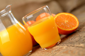 Orange juice in a glass, carafe with fresh juice and fresh oranges in the background on wooden table