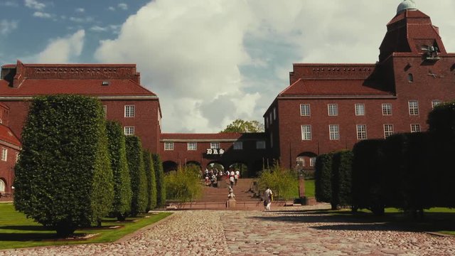 Wide view of a cobblestone road lined by trimmed trees leading to a fountain. Steps behind lead up a hill to brick buildings and an arched entryway as people walk into view. Low angle.