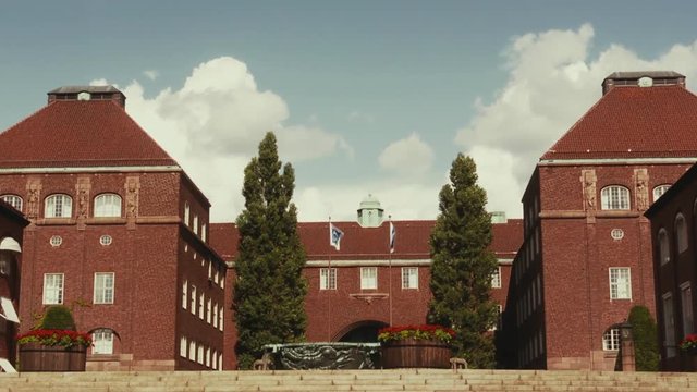 Buildings at the entrance of the KTH Royal Institute of Technology campus. Clean brick buildings around a courtyard with flags flying from tall flag poles. Blue sky and soft clouds.