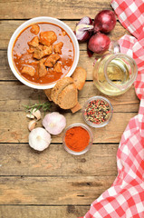 Top view of pork goulash with pieces of meat in a bowl, garlic, pepper, onion, jug with oil and red checkered tablecloth in the background - vertical photo