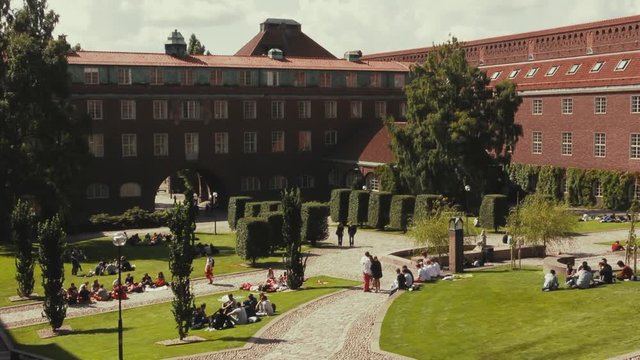 Wide view of students relaxing on the lawn of a courtyard on their university campus. Cobblestone paths and sculpted shrubs and bushes decorate the landscape outside of large dark red brick buildings.