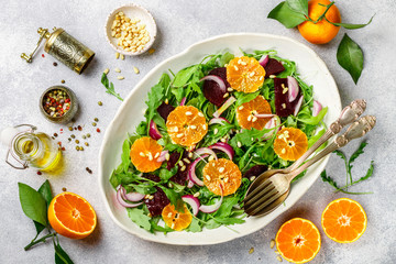 Gourmet salad of arugula, baked beetroot, tangerines and red onions with spices and pine nuts in a white dish. Olive oil and ingredients on a grey surface. Selective focus
