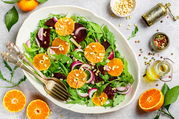 Delicatessen colorful salad of baked beets, arugula, tangerines and red onions with spices and pine nuts in a white dish. Olive oil and ingredients on a grey surface