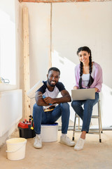 smiling african american couple with laptop and paint roller looking at camera during renovation of home