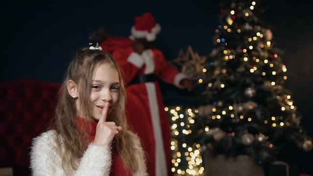 Little cute girl put finger on her lips asking to be quieter while santa claus putting gift.