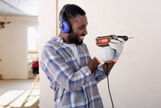 Side View Of Laughing Young African American Man In Protective Headphones And Gloves Having Fun With Power Drill During Renovation Of Home