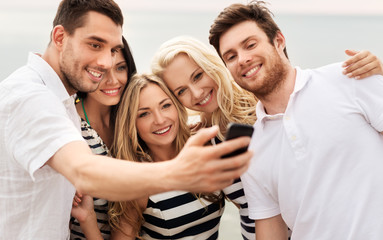 friendship, summer holidays and people concept - group of happy friends taking picture by smartphone on beach