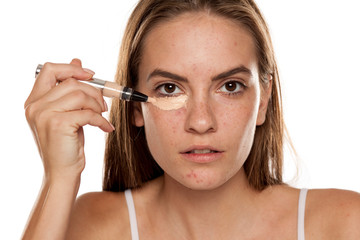 young beautiful girl applying concealer under her eyes on white backgeound