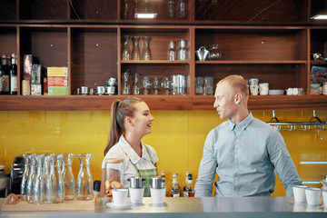 Young man and woman standing at counter of coffee shop and talking while working together