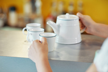 Faceless shot of waiter at counter with white teapot and few cups before serving