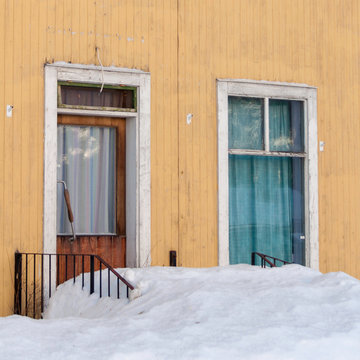 Once a cafe, now neglected and forgotten with snow covering the steps and electrical wires hanging loose on this yellow house with turqouise curtains.
