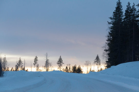 Snowy road leading upwards, opening from dark forest to clear-cut on the top of the hill and sunset, lots of sky.