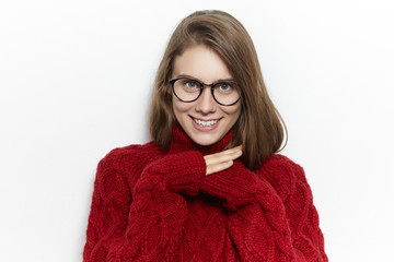 Horizontal image of cute joyful teenage girl wearing maroon turtleneck sweater while spending cold autumn day at home, warming up, hiding hands inside sleeves and smiling happily at camera