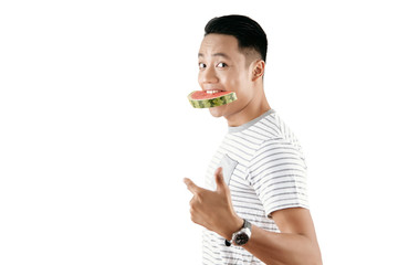 Portrait of young Asian man holding watermelon slice in his mouth and looking at camera against white background, copy space