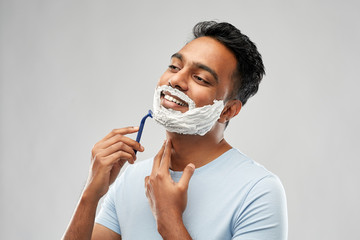 grooming and people concept - young indian man shaving beard with manual razor blade over grey background - Powered by Adobe