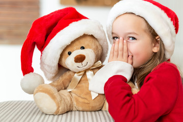 Cute young girl wearing santa hat whispering a secret to her teddy bear christmas present toy. Cheeky kid sharing secrets with teddy bear.