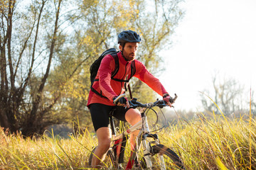 Determined young bearded man in red long sleeve cycling jersey riding mountain bike along a path through tall grass. Low angle front side view.