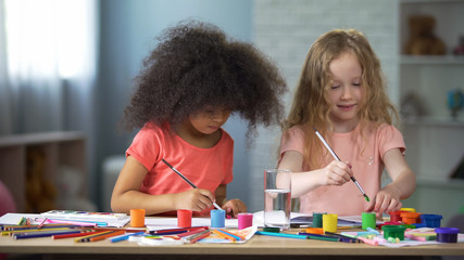 Cheerful African and European girls sitting at the table and painting, hobby