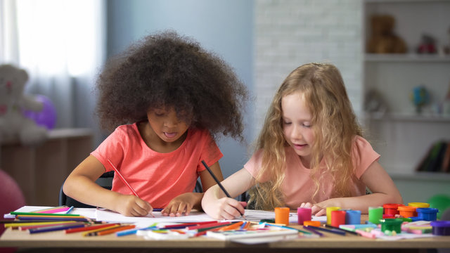 Two multi-ethnic girls drawing at table with colorful pencils, happy childhood