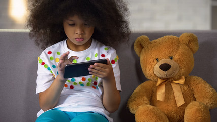 Preschool curly african girl studying letters on smartphone, educational apps