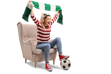 Young femal soccer fan cheering with a scarf and sitting in an armchair