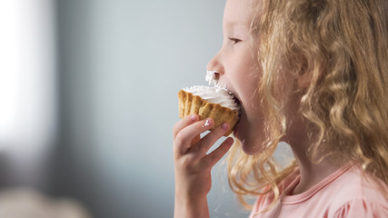 Cute pale preschool girl eating creamy cake and smiling, candy bar at party