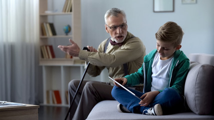 Angry grandfather scolding boy for playing video game on tablet, generation gap - obrazy, fototapety, plakaty