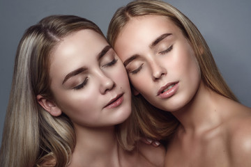 Close up portrait of two beautiful blond girls with natural nude make up and perfect skin posing with closed eyes. Isolated on grey background. Studio shot
