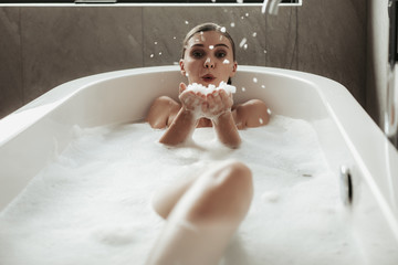 Concept of sensual water care and enjoyment at home. Full length portrait of beautiful brunette woman blowing foam while taking relaxing bath