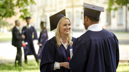 Female graduate in square hat with diploma talking to classmate, convocation day