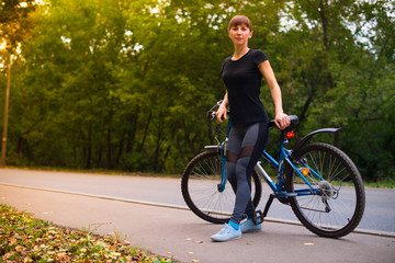 Young beautiful athletic girl in sportswear sitting on a blue bike frame. Trees on the background.