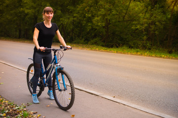 Young beautiful athletic girl in sportswear sitting on a blue bike on the sidewalk. Trees on the background.