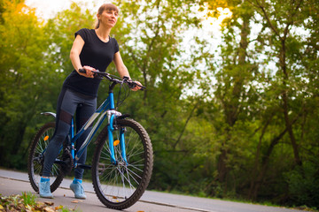 Young beautiful athletic girl in sportswear sitting on a blue bike on the sidewalk. Trees on the background.