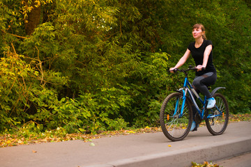 young beautiful girl in sportswear rides a blue bike on the sidewalk. Trees on the background.
