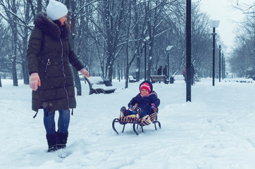 Fototapeta na wymiar Mom pulls daughter on sled in winter park in snowfall