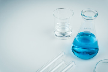laboratory equipments. A blue liquid flask, clear liquid beaker, test tubes and petri dish on the white table