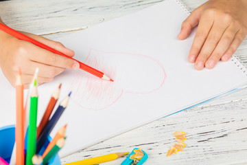 Hand of children drawing red heart with colored pencil on white paper on wooden table.