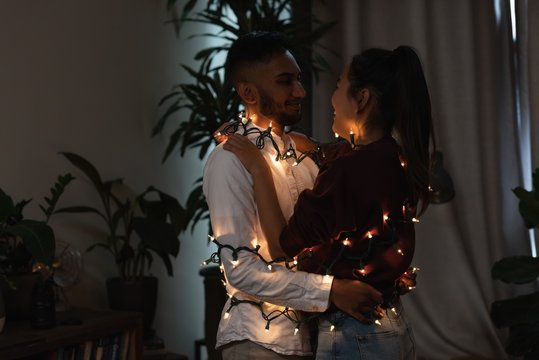 Couple wrapped in fairy light embracing each other at home