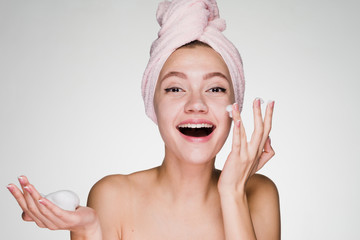 happy young girl with a pink towel on her head and white teeth apply cleansing foam on her face