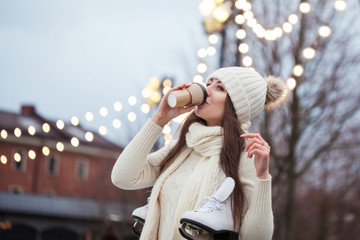 Happy young woman in knitted sweater and hat is going skating and drinks coffee